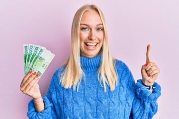 Young blonde girl holding 10000 south korean won banknotes smiling with an idea or question pointing finger with happy face, number one