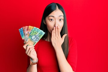 Young chinese woman holding australian dollars covering mouth with hand, shocked and afraid for mistake. surprised expression