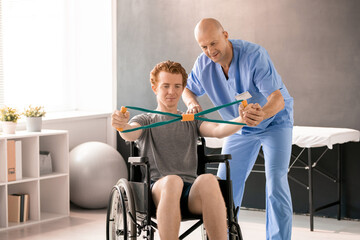 Fototapeta na wymiar Mature clinician in blue uniform supporting hand of young patient in wheelchair