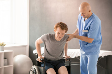 Fototapeta na wymiar Mature clinician in uniform helping young man in pain to sit in wheelchair