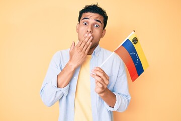 Young handsome hispanic man holding venezuelan flag covering mouth with hand, shocked and afraid...