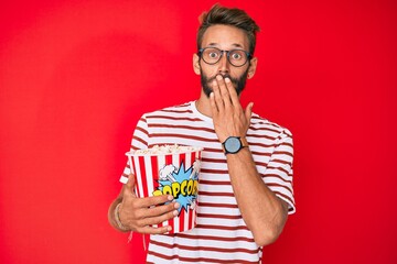 Handsome caucasian man with beard eating popcorn covering mouth with hand, shocked and afraid for...