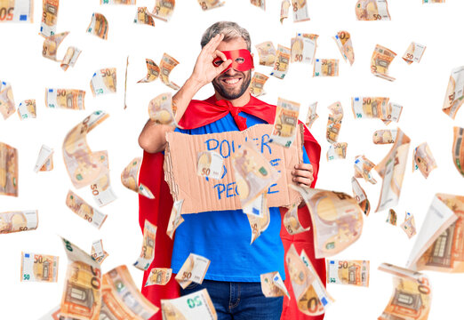 Young blond man wearing super hero custome holding power to the people cardboard banner smiling happy doing ok sign with hand on eye looking through fingers
