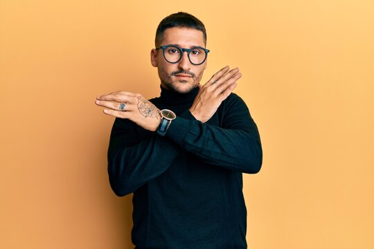 Handsome man with tattoos wearing turtleneck sweater and glasses rejection expression crossing arms doing negative sign, angry face