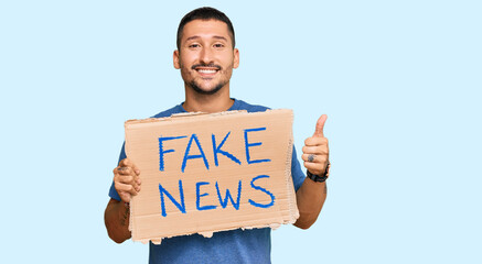 Handsome man with tattoos holding fake news banner smiling happy and positive, thumb up doing excellent and approval sign