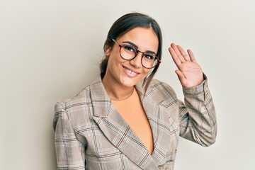 Young brunette woman wearing business jacket and glasses waiving saying hello happy and smiling, friendly welcome gesture