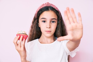 Cute hispanic child girl holding cupcake with open hand doing stop sign with serious and confident...