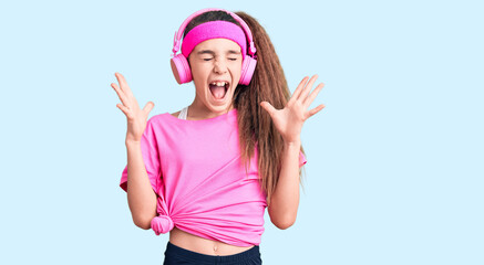 Obraz na płótnie Canvas Cute hispanic child girl wearing gym clothes and using headphones celebrating mad and crazy for success with arms raised and closed eyes screaming excited. winner concept