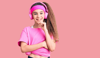 Obraz na płótnie Canvas Cute hispanic child girl wearing gym clothes and using headphones looking confident at the camera with smile with crossed arms and hand raised on chin. thinking positive.