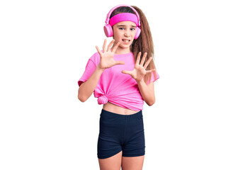Cute hispanic child girl wearing gym clothes and using headphones disgusted expression, displeased...