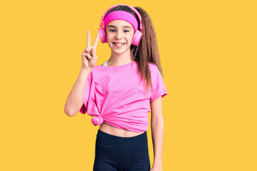 Cute hispanic child girl wearing gym clothes and using headphones showing and pointing up with fingers number two while smiling confident and happy.