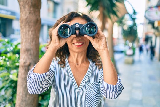 Middle age hispanic woman smiling happy looking for new opportunity using binoculars at the city.