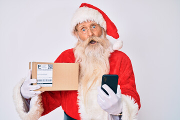 Old senior man with grey hair and long beard wearing santa claus costume holding delivery box and phone in shock face, looking skeptical and sarcastic, surprised with open mouth