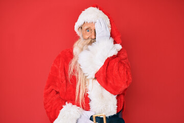 Old senior man with grey hair and long beard wearing traditional santa claus costume covering one eye with hand, confident smile on face and surprise emotion.