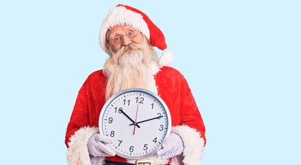 Old senior man with grey hair and long beard wearing santa claus costume holding clock looking positive and happy standing and smiling with a confident smile showing teeth