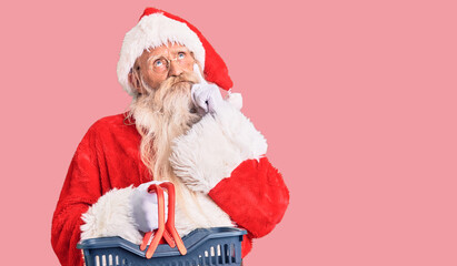 Old senior man with grey hair and long beard wearing santa claus costume holding shopping basket serious face thinking about question with hand on chin, thoughtful about confusing idea