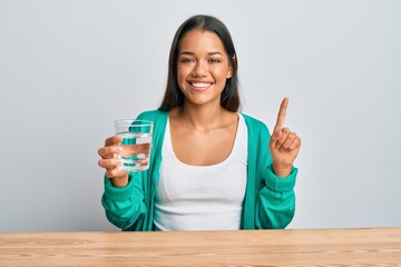 Beautiful hispanic woman drinking glass of water smiling with an idea or question pointing finger with happy face, number one