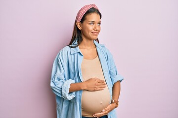 Beautiful hispanic woman expecting a baby, touching pregnant belly smiling looking to the side and...
