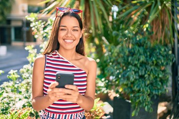 Young latin girl smiling happy using smartphone at the city.