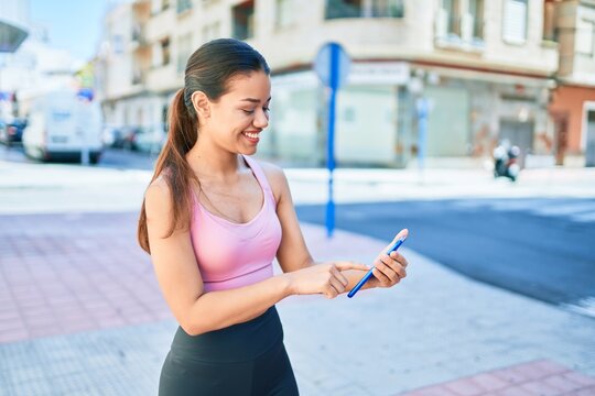 Young beautiful hispanic sporty woman wearing fitness outfit smiling happy and natural texting using smartphone at the town
