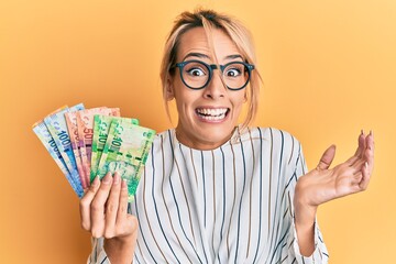 Beautiful blonde woman holding south african rand banknotes celebrating achievement with happy...
