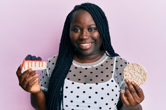 Young Black Woman With Braids Eating Cake Slice And Rice Crackers Smiling With A Happy And Cool Smile On Face. Showing Teeth.