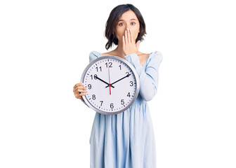 Young beautiful girl holding big clock covering mouth with hand, shocked and afraid for mistake....