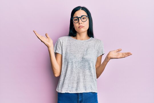 Beautiful young woman wearing casual clothes and glasses clueless and confused with open arms, no idea and doubtful face.
