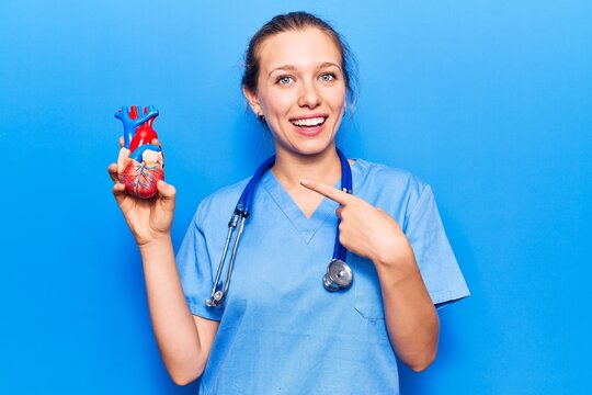 Young blonde woman wearing doctor uniform holding heart smiling happy pointing with hand and finger