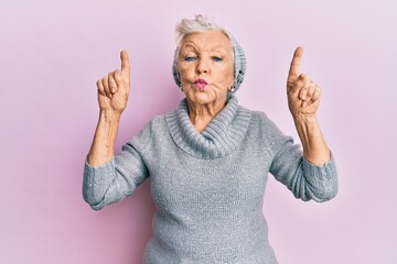 Senior grey-haired woman pointing up with fingers looking at the camera blowing a kiss being lovely...