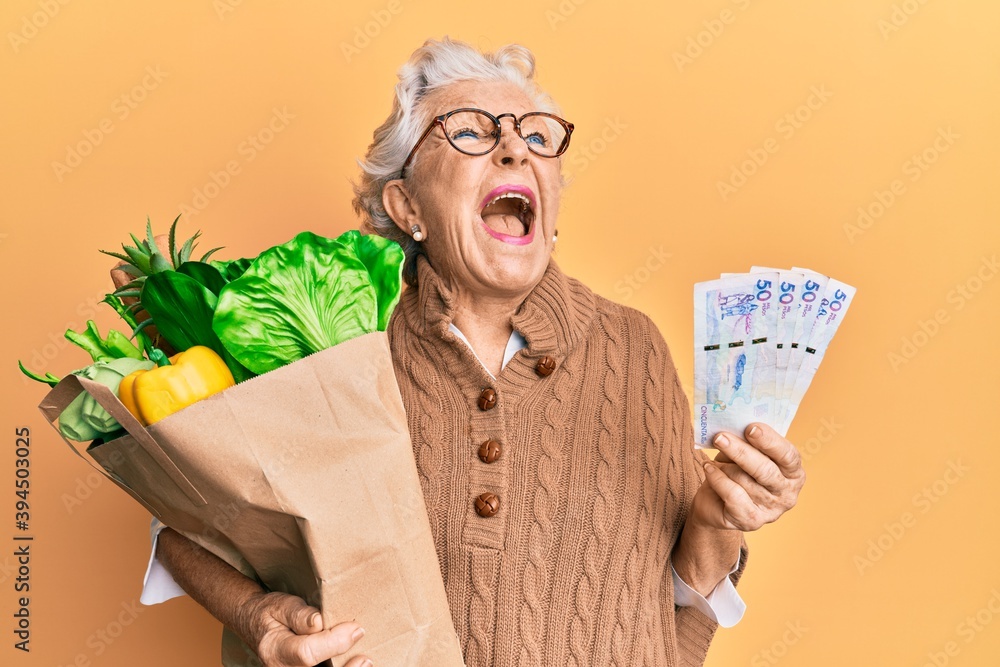 Sticker senior grey-haired woman holding groceries and colombian pesos banknotes angry and mad screaming fru