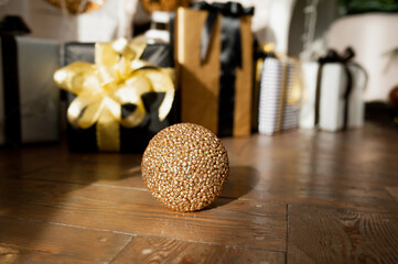round Golden shining ball rolling on wooden parquet floor with holiday gift boxes on background in natural sunlight, selective focus