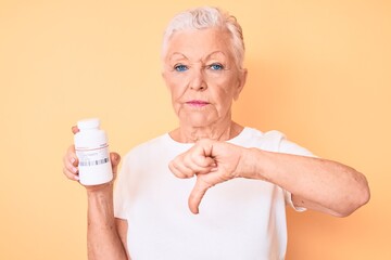 Senior beautiful woman with blue eyes and grey hair holding pills with angry face, negative sign showing dislike with thumbs down, rejection concept