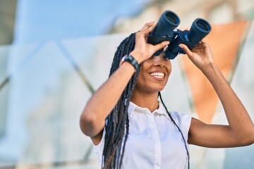 Young african american woman smiling happy looking for new opportunity using binoculars at the city.