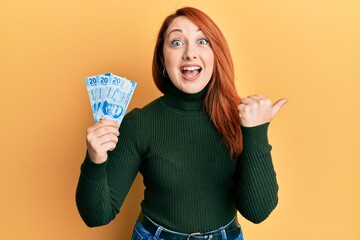 Beautiful redhead woman holding 20 mexican pesos banknotes pointing thumb up to the side smiling happy with open mouth