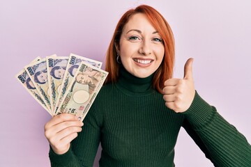 Beautiful redhead woman holding japanese yen banknotes smiling happy and positive, thumb up doing excellent and approval sign