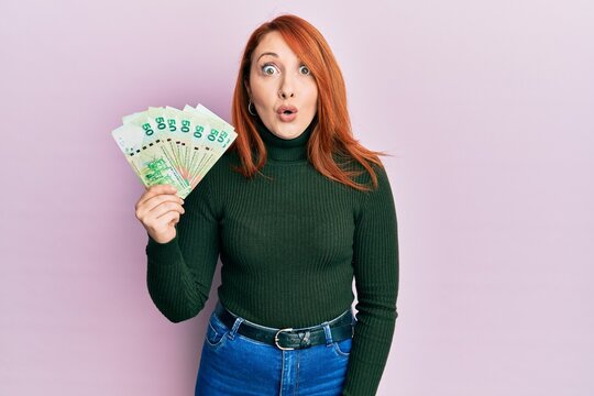 Beautiful redhead woman holding 50 hong kong dollars banknotes scared and amazed with open mouth for surprise, disbelief face