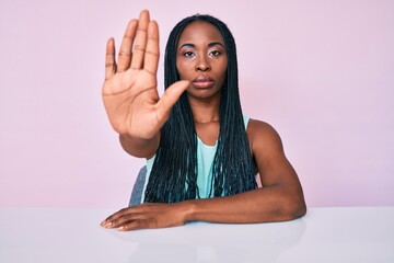 African american woman with braids wearing casual clothes sitting on the table doing stop sing with...