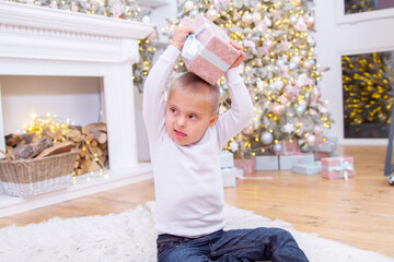 A boy with Down syndrome and genetic pathology sits by the New Year tree and holds a box with a...
