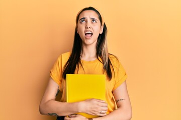 Young hispanic woman holding book winking looking at the camera with sexy expression, cheerful and...