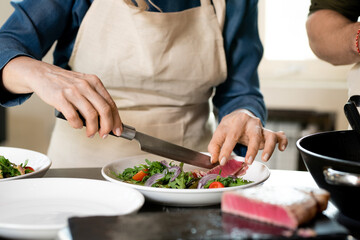 Hands of mature female putting slice of smoked meat on plate with salad