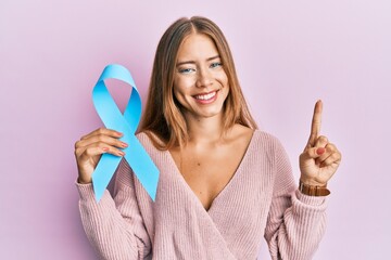 Beautiful young blonde woman holding blue ribbon smiling with an idea or question pointing finger...