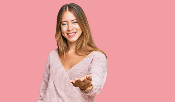 Beautiful blonde woman wearing casual winter pink sweater smiling cheerful offering palm hand giving assistance and acceptance.