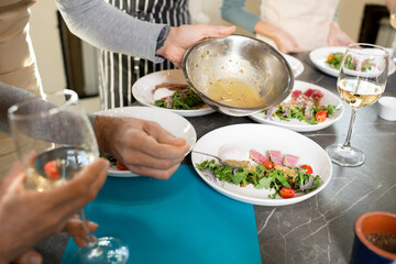 Hands of mature man putting sauce from metallic bowl on plate with salad