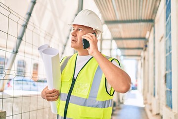 Middle age architect man talking on the smartphone and holding blueprints at the city.