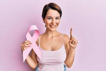 Young brunette woman with short hair holding pink cancer ribbon smiling with an idea or question pointing finger with happy face, number one