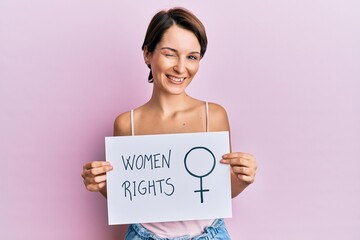 Young brunette woman with short hair holding woman rights banner winking looking at the camera with...