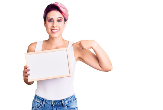 Young beautiful woman with pink hair holding empty white chalkboard pointing finger to one self smiling happy and proud