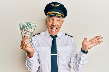 Handsome middle age mature man wearing airplane pilot uniform holding dollars celebrating...