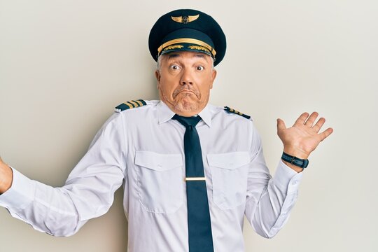 Handsome middle age mature man wearing airplane pilot uniform clueless and confused expression with arms and hands raised. doubt concept.